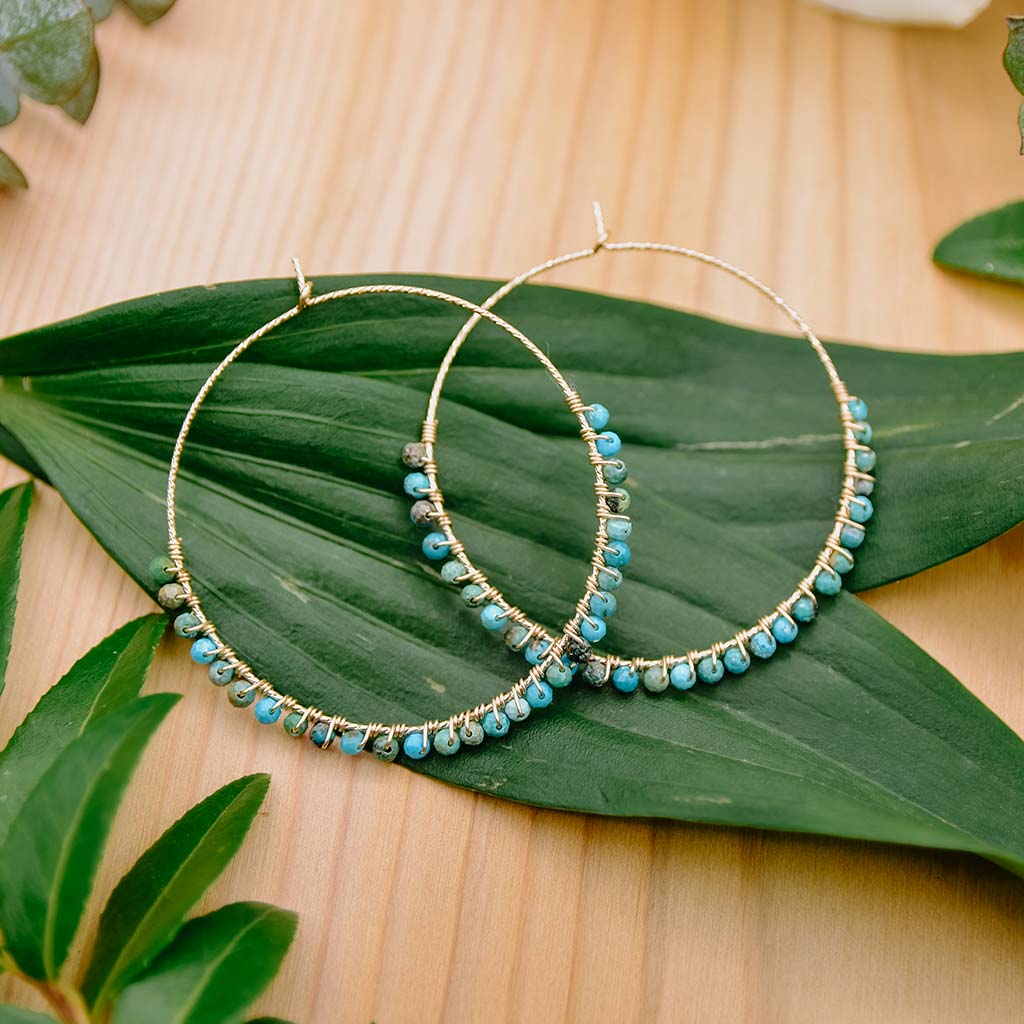 This image show a pair of 14k hoops with tiny turquoise blue gemstones wire wrapped on the hoops. Earrings and other natural gemstones jewelry by Emerald Sun Creations