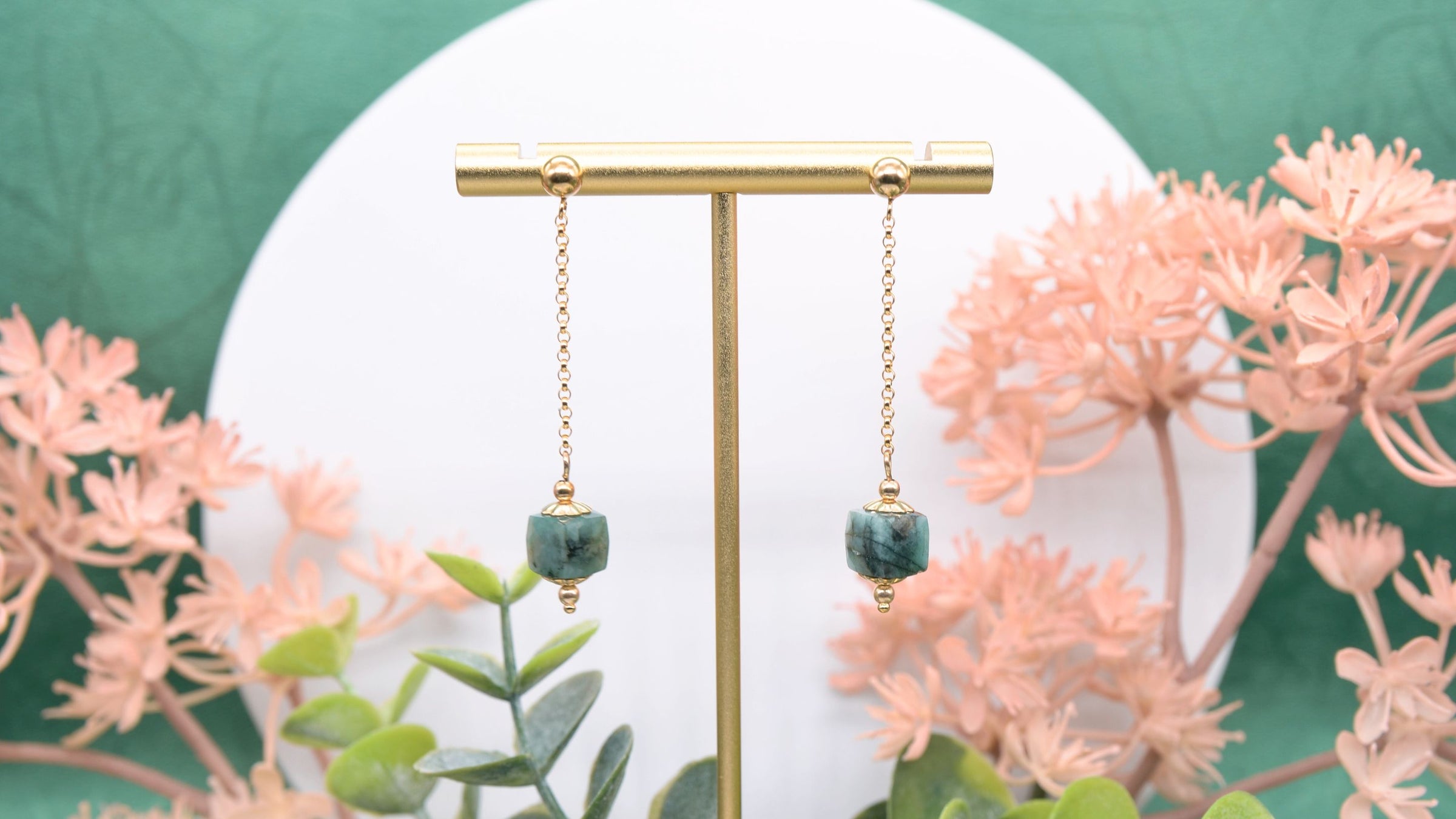 The image shows a pair of earrings with Emerald gemstones and gold chain with greenery surrounding it. Included are 2 buttons that say "Shop All" on the left and "All Earrings" on the right. Natural gemstone jewelry by Emerald Sun Creations