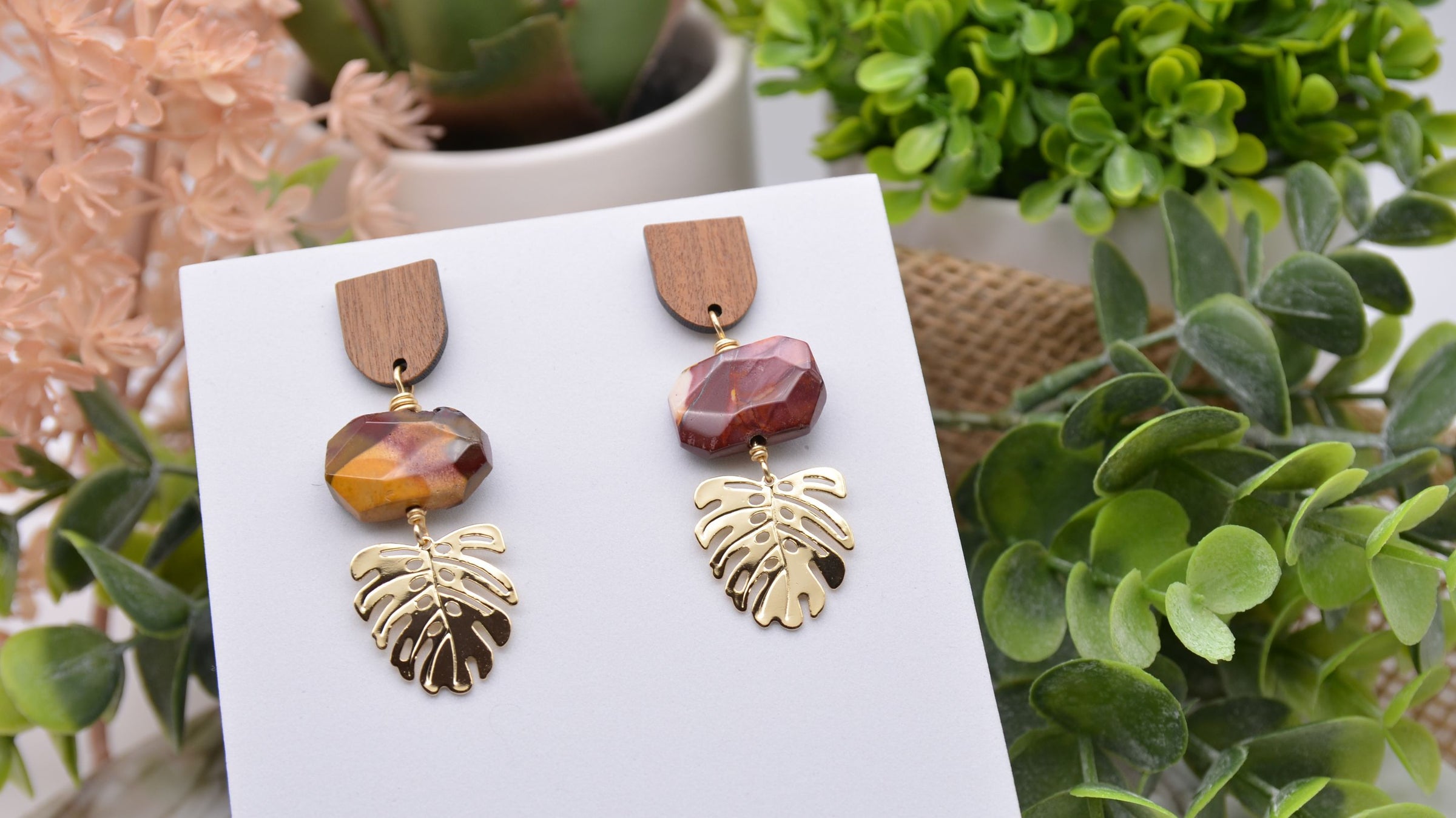 The image shows a pair of earrings with wooden semicircle ear post, orange and maroon colored Mookaite gemstones and gold leaf charms on a white display with greenery surrounding it. The words "New In Earrings, Fall Hues, Transition into Fall like a pro" at the bottom. 2 buttons that say "Shop All" on the left and "All Earrings" on the right. Natural gemstone jewelry by Emerald Sun Creations