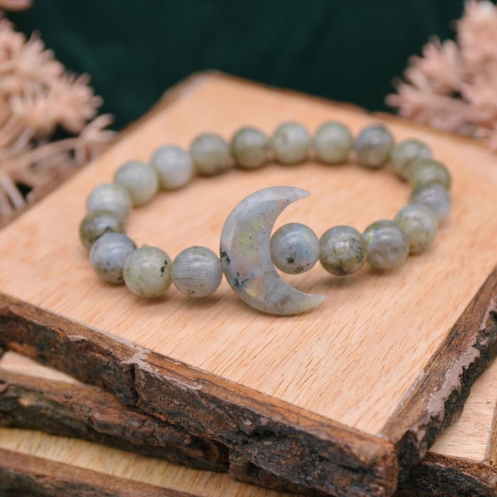 A labradorite gemstone bracelet with a crystal crescent moon and labradorite beads displayed on a wooden background. By Emerald Sun Creations