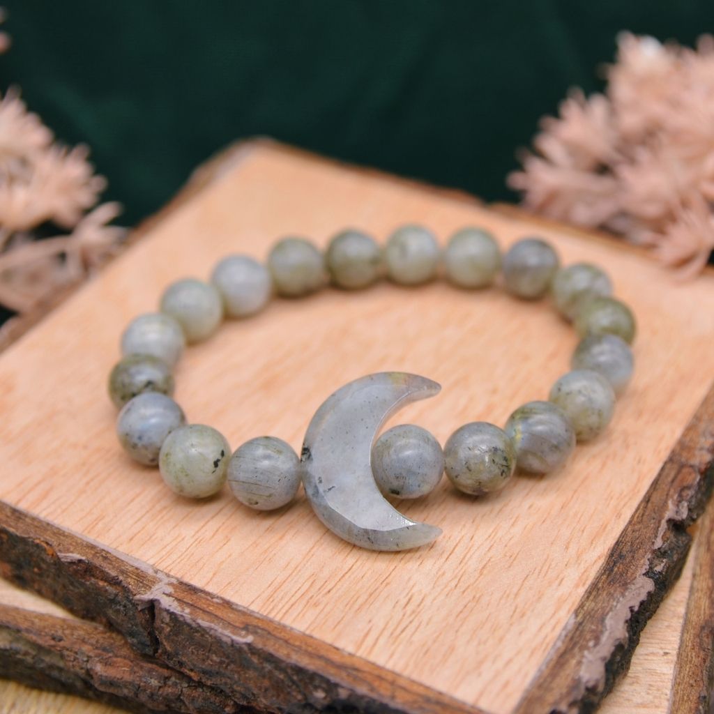 A labradorite gemstone bracelet with a crystal crescent moon and labradorite beads displayed on a wooden background. By Emerald Sun Creations