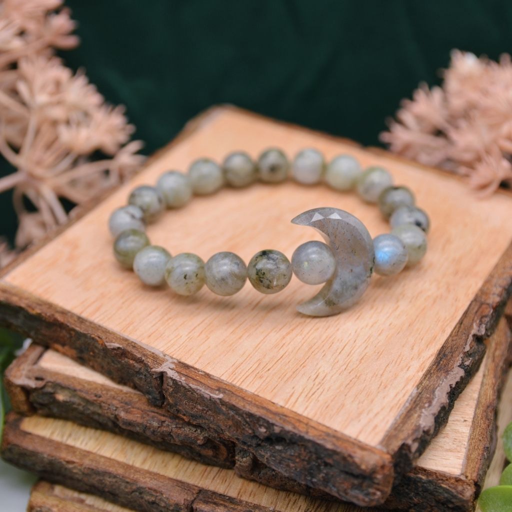 A labradorite gemstone bracelet with a crystal crescent moon and labradorite beads displayed on a wooden background. By Emerald Sun Creations