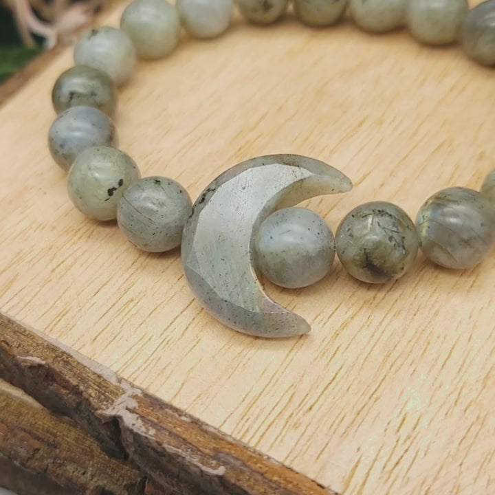 A video of a labradorite gemstone bracelet with a crystal crescent moon and labradorite beads displayed on a wooden background. By Emerald Sun Creations